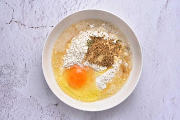 A bowl with an egg, flour, spices, and liquid ingredients for beer battered chicken tenders sits ready to be mixed on a light surface.