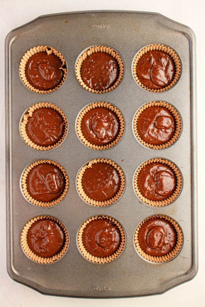 A muffin pan holds twelve chocolate cupcake liners brimming with rich brownie mix, ready to transform into delectable brownie cupcakes in the oven.