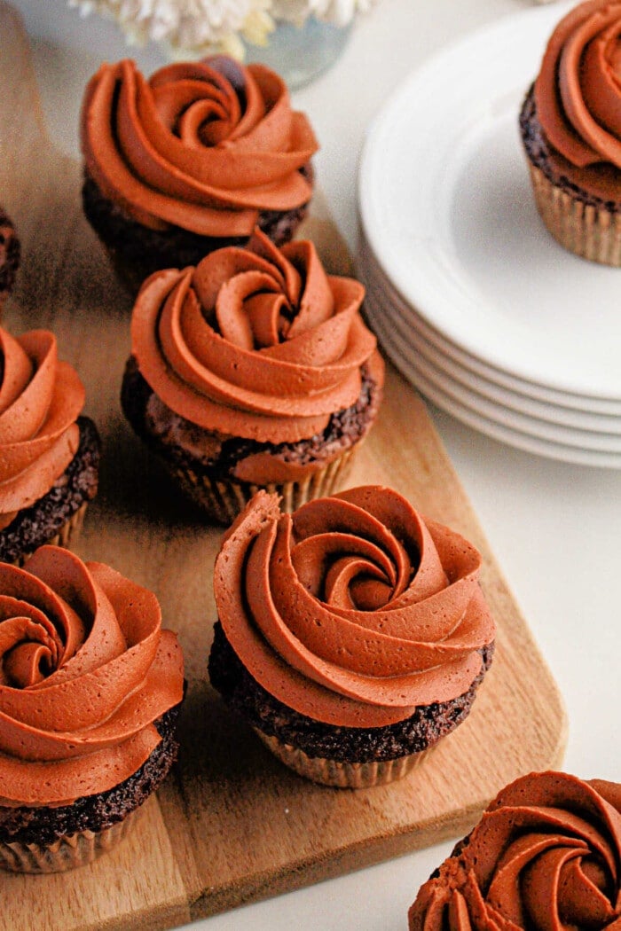 Brownie cupcakes with swirl frosting sit elegantly on a wooden board next to stacked white plates.