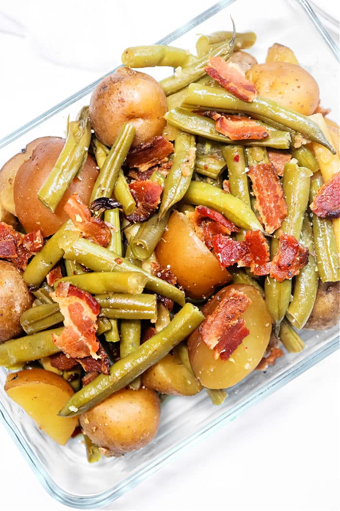 Green Beans with Ham and Potatoes are served together in a clear glass dish, showcasing green beans, small potatoes, and savory bacon pieces on a white background.