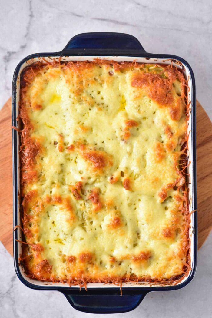 Baked cheesy pasta casserole made with baked frozen ravioli, featuring a golden, bubbly top in a rectangular blue baking dish on a wooden board.