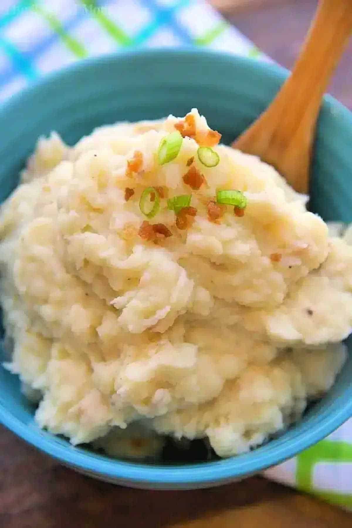 A bowl of pressure cooker mashed potatoes topped with chopped green onions and bacon bits, with a wooden spoon in it.