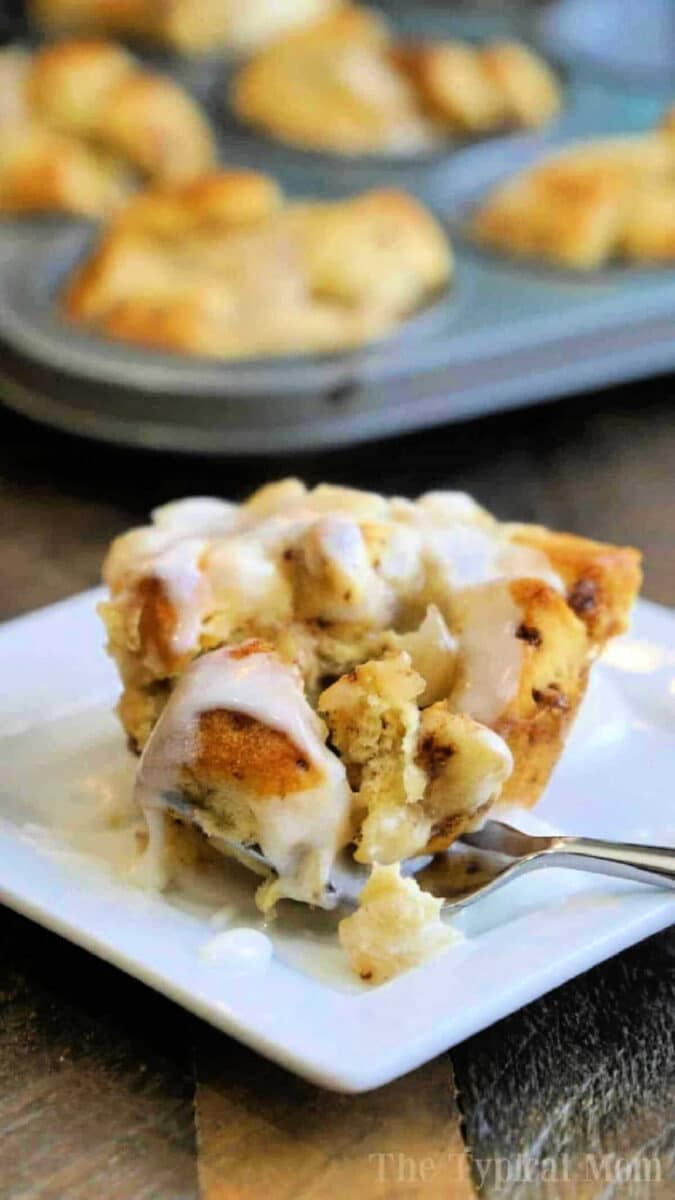 A gooey cinnamon roll muffin with icing on a white plate, with more monkey bread muffins in a tray in the background.