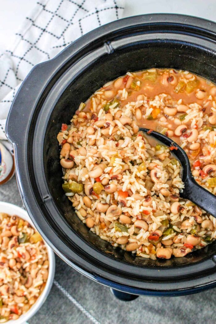 A slow cooker filled with Black Eyed Peas and Rice, diced vegetables, and a black serving spoon.