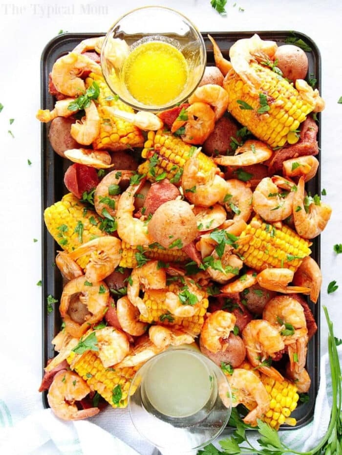 A tray of instant pot shrimp boil with corn, potatoes, sausage, herbs, and two bowls of dipping sauces.