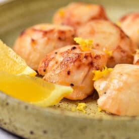 Seared scallops garnished with lemon zest, served with lemon wedges on a green ceramic plate&mdash;perfectly cooked once you know how long to cook scallops for tender, golden results.