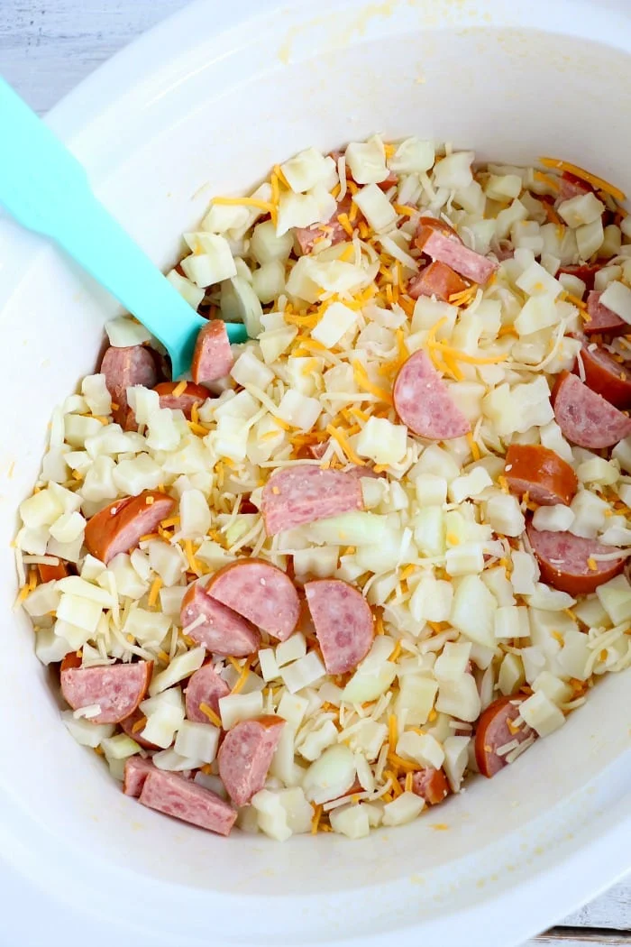 Chopped sausage, slow cooker cheesy potatoes, and shredded cheese are mixed together in a white slow cooker with a blue spatula.