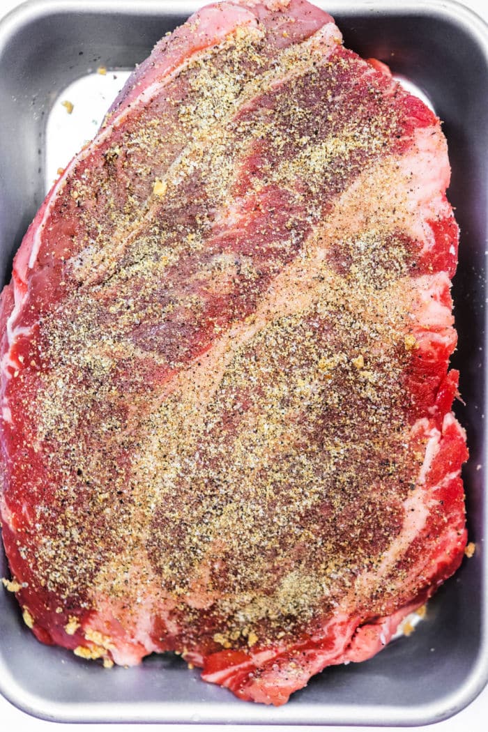 Raw seasoned beef brisket in a metal baking pan, coated with pepper and spices, ready for cooking&mdash;perfect for those who love the flavors of a classic smoked chuck roast.
