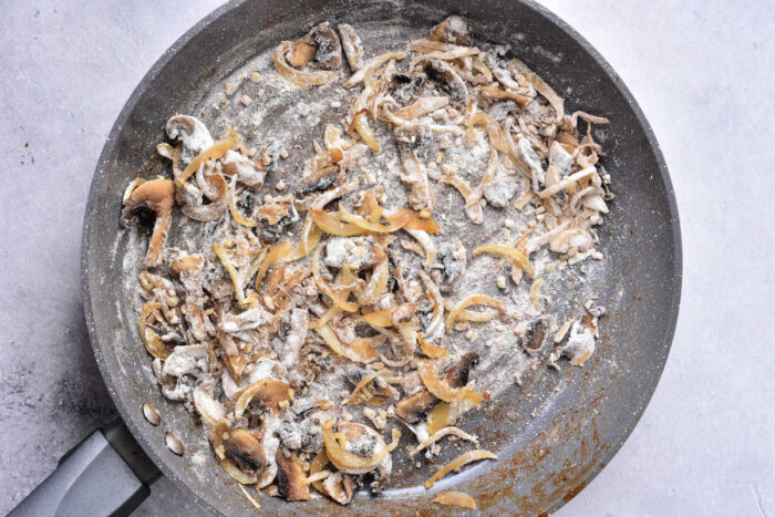 Sliced mushrooms and onions cooking with flour in a large skillet&mdash;an essential step in preparing hearty Venison Stroganoff&mdash;on a light countertop.