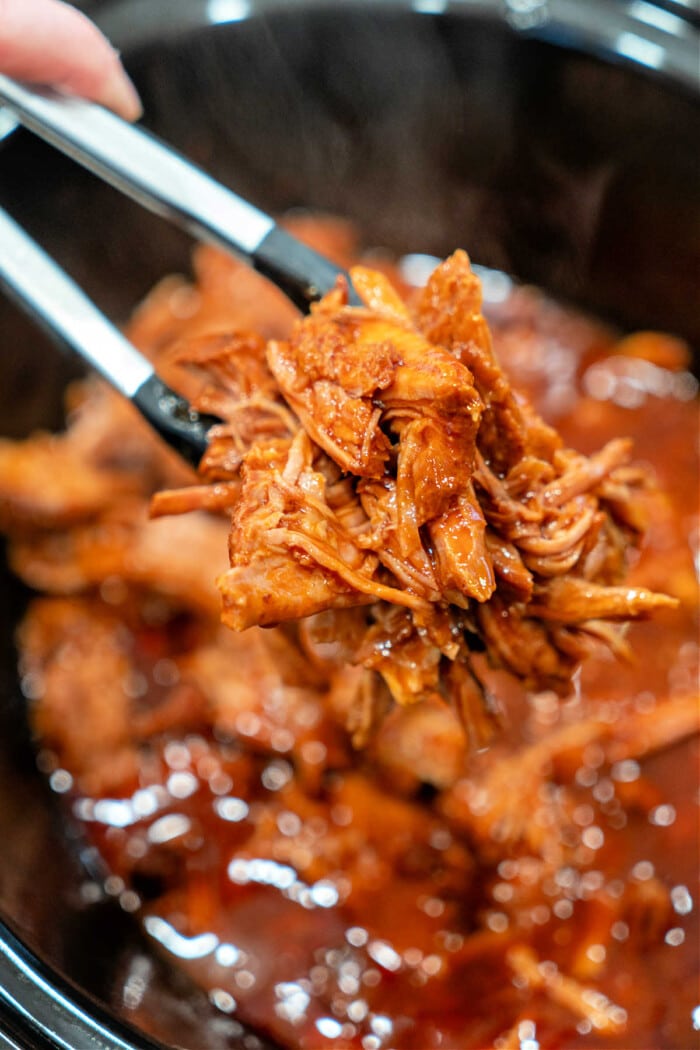 Tongs holding shredded BBQ pulled pork over a slow cooker full of this mouthwatering 2 ingredient pulled pork recipe.