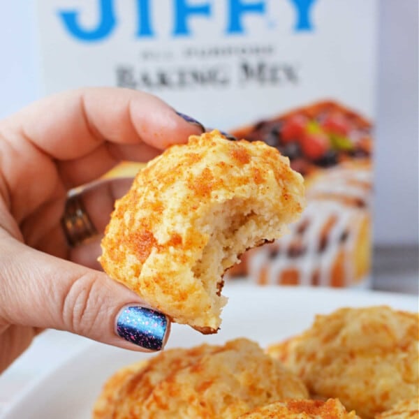 A hand holds a bitten cheddar biscuit, with a Jiffy Mix box adding a tantalizing promise of more flaky treats in the background.