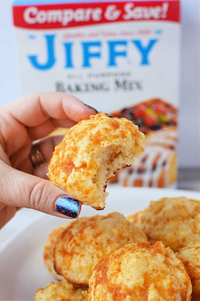 A hand holds a bitten cheddar biscuit, with a Jiffy Mix box adding a tantalizing promise of more flaky treats in the background.