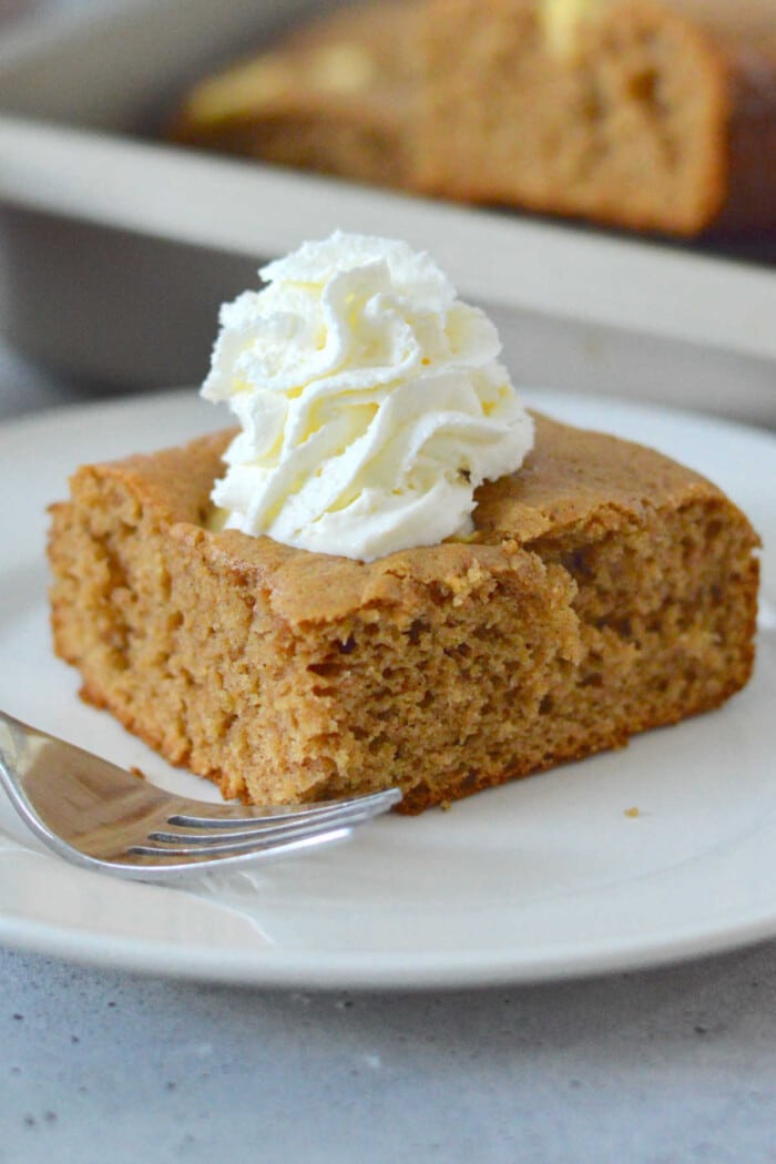 A slice of brown apple cake made with cake mix is topped with whipped cream on a white plate, with a fork beside it.