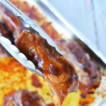 Tongs holding a piece of beautifully glazed boneless pork ribs above a baking tray, ready for the oven, with more succulent, glazed meat waiting beneath.