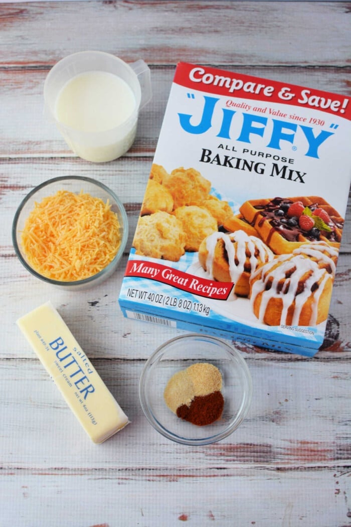 A rustic spread featuring Jiffy Mix biscuits paired with milk, shredded cheese, butter, and spices on a wooden surface.