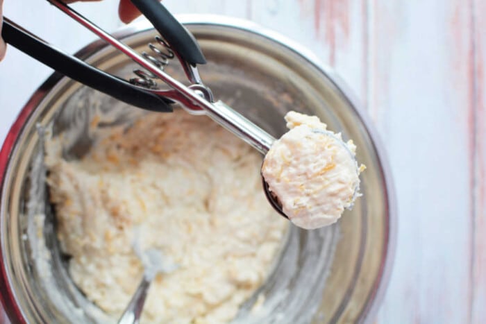 A cookie dough scoop filled with Jiffy Mix dough is held over a mixing bowl on a wooden surface, ready to bake delicious biscuits.