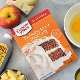 An array of tempting ingredients awaits on the kitchen counter: a box for Apple Cake with Cake Mix, creamy pudding, aromatic cinnamon, chopped apples, an egg, golden syrup, and a trusty measuring cup to tie it all together.