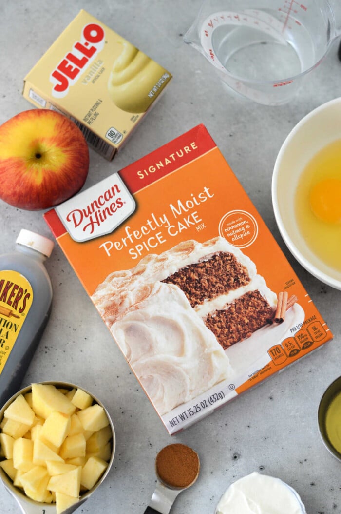 An array of tempting ingredients awaits on the kitchen counter: a box for Apple Cake with Cake Mix, creamy pudding, aromatic cinnamon, chopped apples, an egg, golden syrup, and a trusty measuring cup to tie it all together.