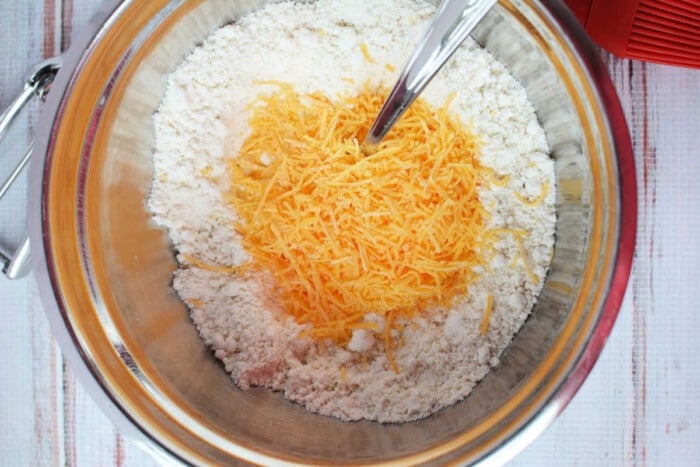 A mixing bowl filled with flour, shredded cheddar cheese, and a spoon rests on a wooden surface, resembling the perfect start for savory biscuits using Jiffy Mix.