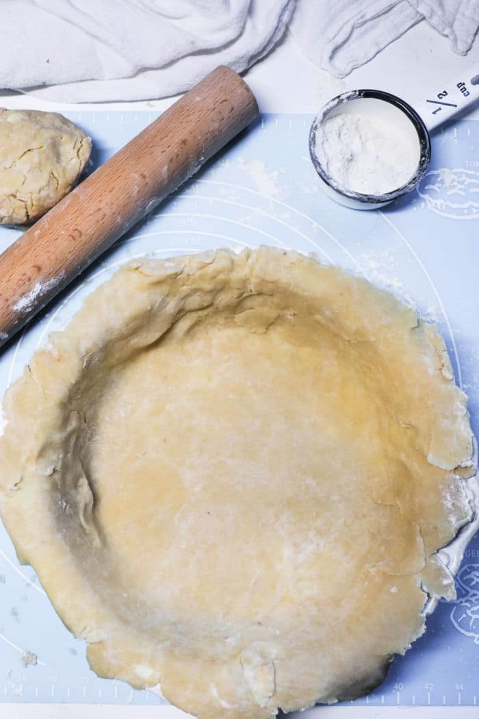 Unbaked pie crust in a pan, with a rolling pin, dough ball, and cup of flour nearby on a blue surface&mdash;perfect foundations for a homemade Double Crust Pot Pie.