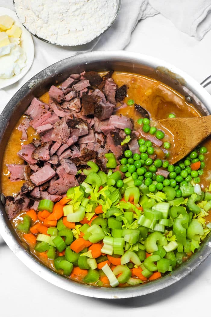 A skillet with diced meat, peas, chopped celery, carrots, and creamy sauce being stirred with a wooden spoon&mdash;the perfect filling for a comforting Double Crust Pot Pie.