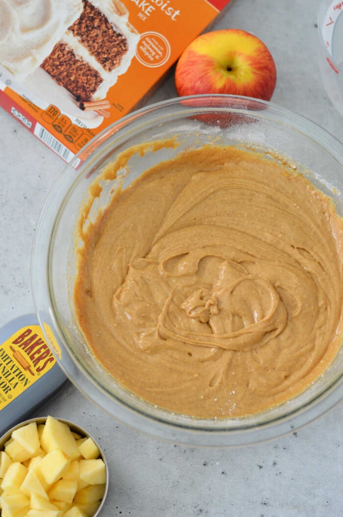 A glass bowl filled with cake batter sits gracefully on the countertop, surrounded by a box of cake mix, chopped apples, a whole apple, and a measuring cup—perfect ingredients for an effortless apple cake.