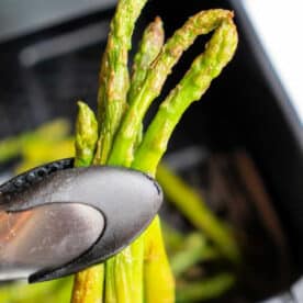 Metal tongs holding cooked asparagus spears above an air fryer basket—discover how to cook frozen asparagus perfectly crisp with this simple air fryer method.