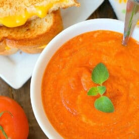 Bowl of creamy tomato soup with herbs, next to a grilled cheese sandwich and fresh tomatoes on a wooden table.