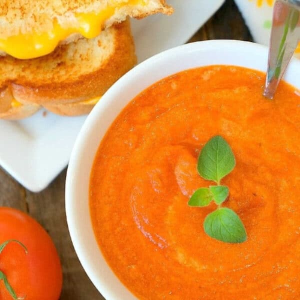 Bowl of creamy tomato soup with herbs, next to a grilled cheese sandwich and fresh tomatoes on a wooden table.