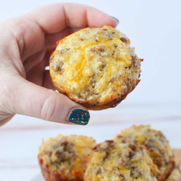 A hand holds a savory muffin crowned with melted cheese, its golden crust hinting at hidden delights within. In the background, a plate overflows with more enticing muffins, each potentially featuring hints of sausage for an added hearty twist. Perfect for those Bisquick-inspired morning feasts!.