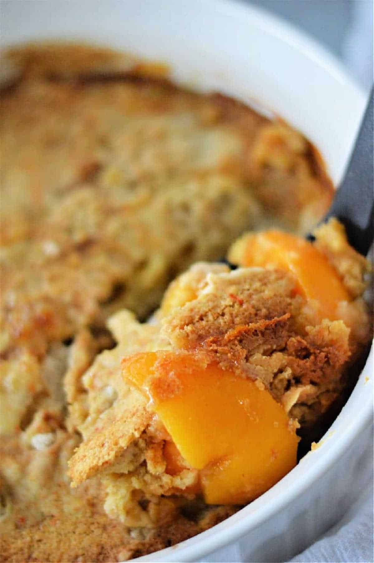 A close-up of a serving of peach cobbler with cake mix being scooped from a white baking dish.