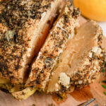 A whole cauliflower head with herbs, perfectly roasted and sliced on a wooden board, sits next to a lemon and fork. This delightful cauliflower recipe is as visually appealing as it is delicious.