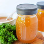 Two mason jars of golden bone broth sit on a wooden board, accompanied by fresh parsley, with a white bowl in the background. Perfect for your favorite slow cooker broth recipe.