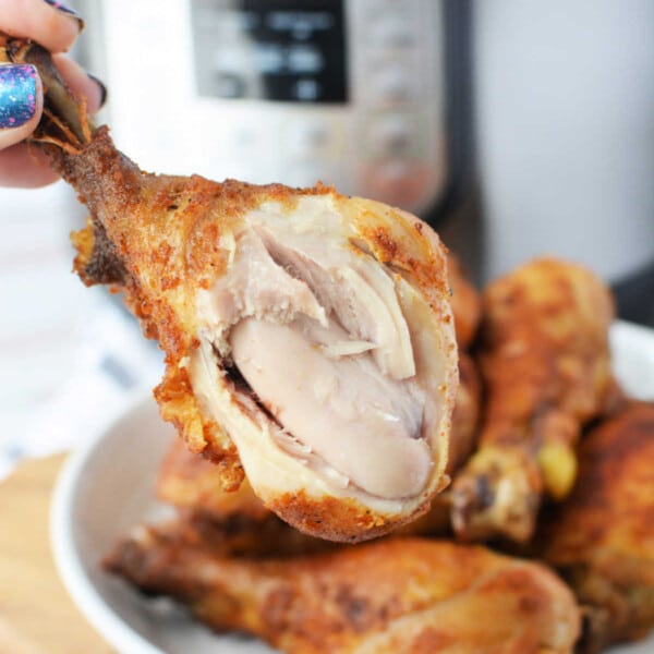 Close-up of a hand holding a juicy piece of pressure cooker fried chicken with visible meat, set against the backdrop of a gleaming pressure cooker.