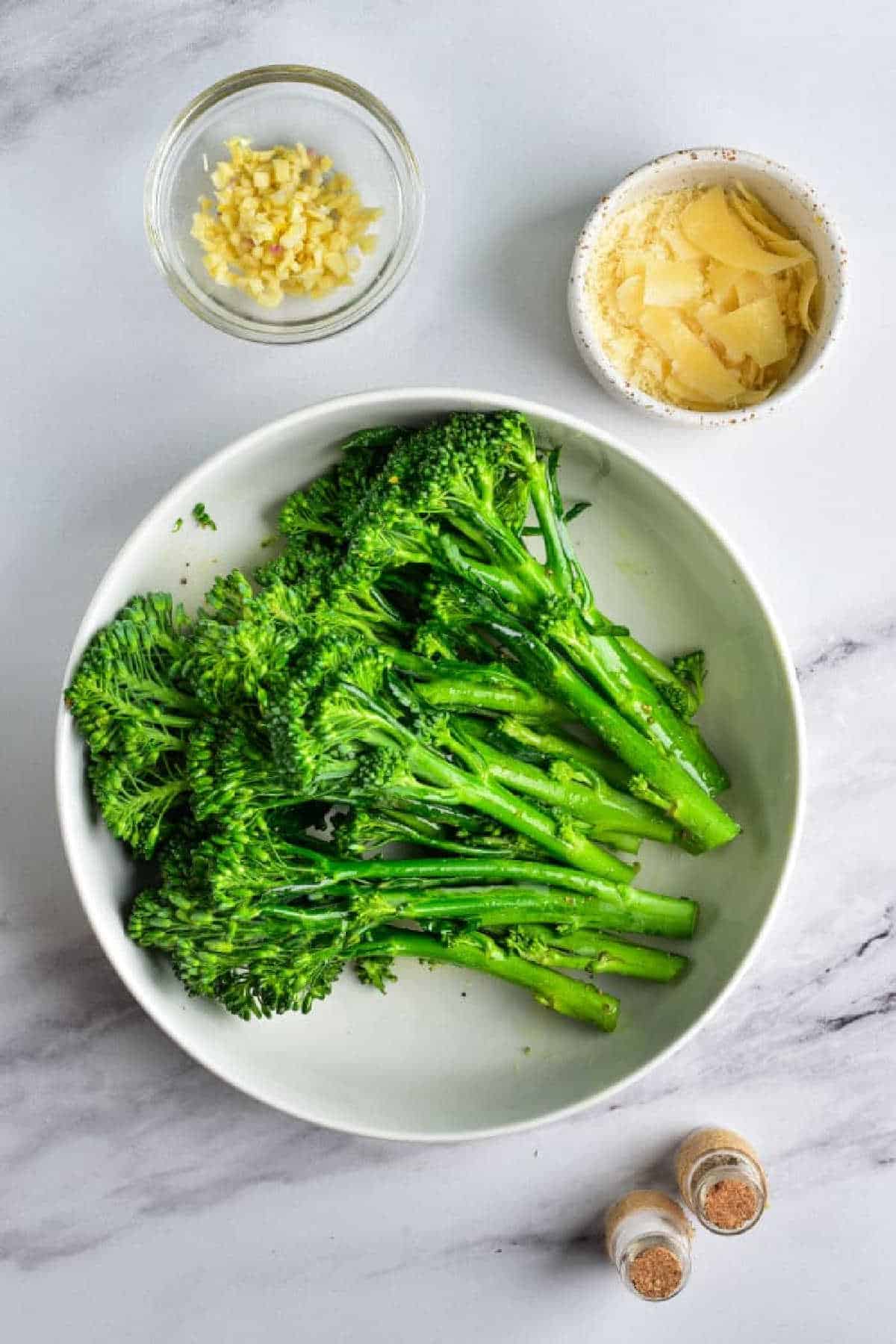 A bowl of Air Fryer Broccolini, with bowls of chopped garlic and parmesan, plus two spice jars on a marble surface.