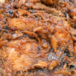 A close-up of braised beef brisket with caramelized onions in a rich, savory roast sauce.