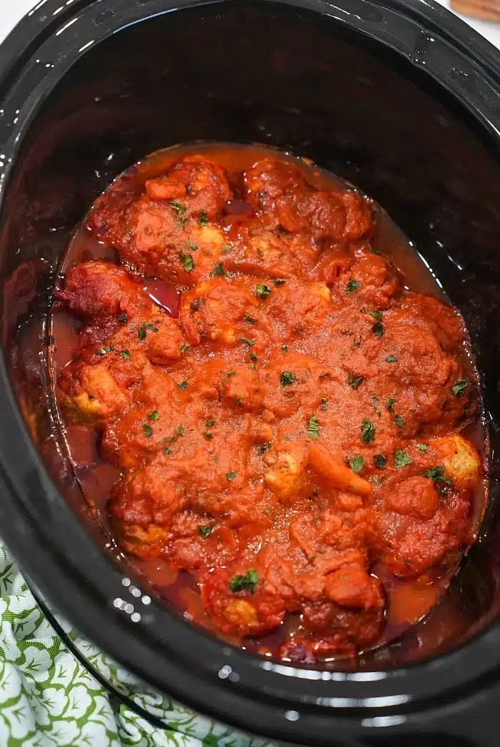 Slow cooker filled with frozen meatballs in crockpot, simmering in tomato sauce and garnished with chopped herbs.