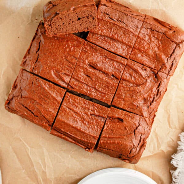 A rich, sliced brownies sits invitingly on parchment paper, a white plate poised nearby, with a hint of greenery in the corner. Try this as a delightful twist on your favorite protein brownie recipe.