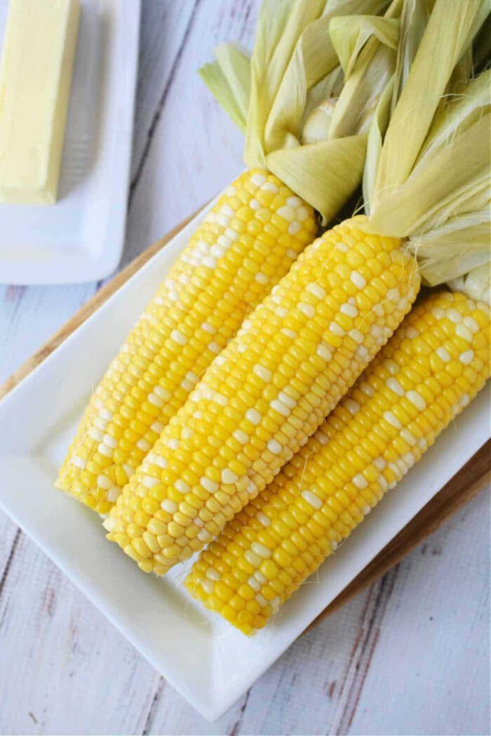 Four ears of fresh corn on the cob with husks, placed on a white rectangular plate—perfect for learning how to cook corn on the cob in the oven for a simple and delicious side dish.