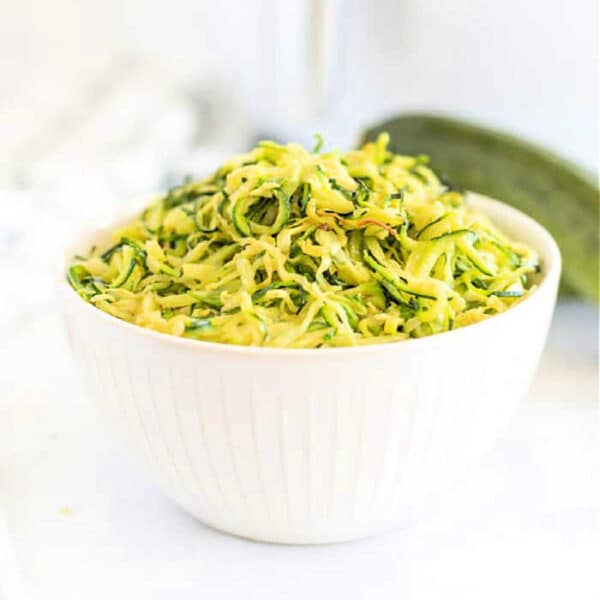 White bowl filled with shredded zucchini on a white surface, perfect for making air fryer zucchini noodles, with a fresh zucchini in the background.