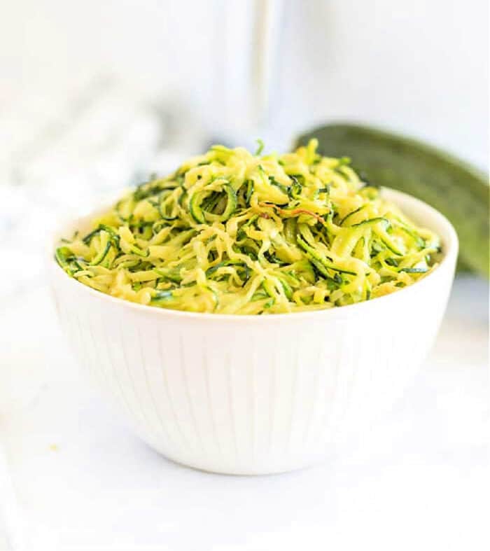 White bowl filled with shredded zucchini on a white surface, perfect for making air fryer zucchini noodles, with a fresh zucchini in the background.