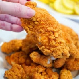 A hand holding a piece of crispy fried chicken above a plate piled with more golden pieces, just like the results you get from cooking frozen chicken tenders in an air fryer.