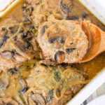 Hamburger steaks in mushroom sauce in a white baking dish, reminiscent of classic Hamburger Steak flavors, with a wooden spoon lifting one piece.