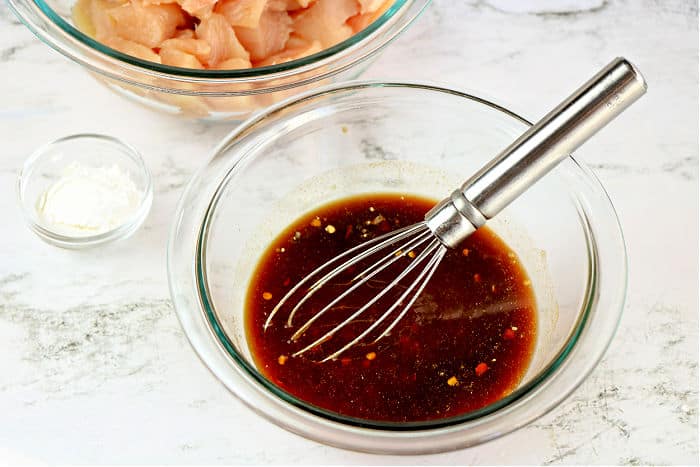 Glass bowl with dark sauce and a whisk, perfect for marinating asian chicken, sits next to a bowl of raw chicken and a small dish of white powder.