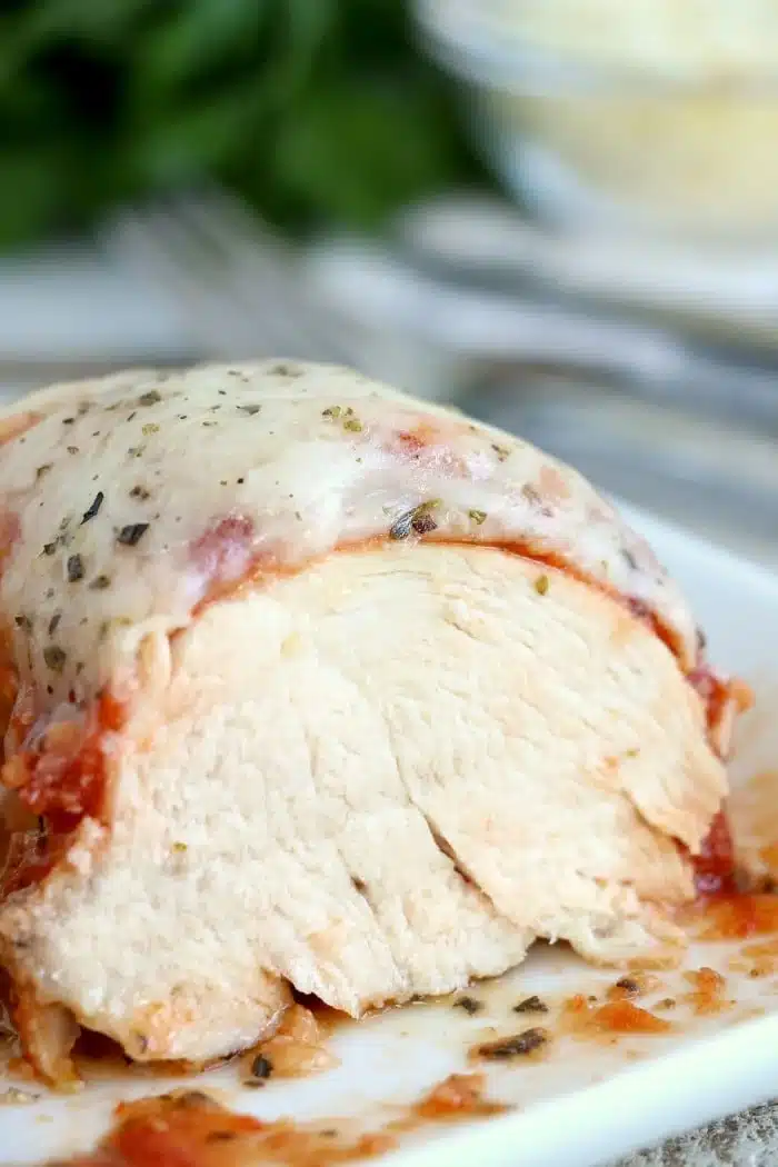 A close-up of instant pot chicken parmesan: sliced, baked chicken breast topped with melted cheese, tomato sauce, and herbs on a plate.