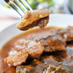 A fork holds a bite of tender meat above a plate of saucy, seasoned pork steak, showcasing a mouthwatering pork steak recipe.