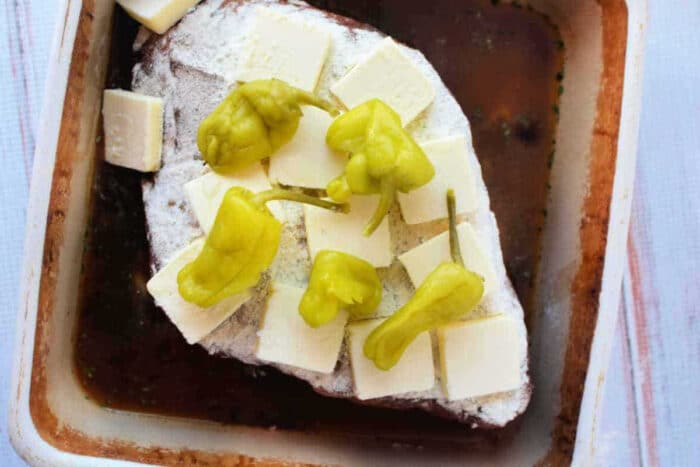 A Mississippi Pot Roast Recipe featuring a roast topped with butter slices and pepperoncini peppers, nestled in a baking dish with savory broth.