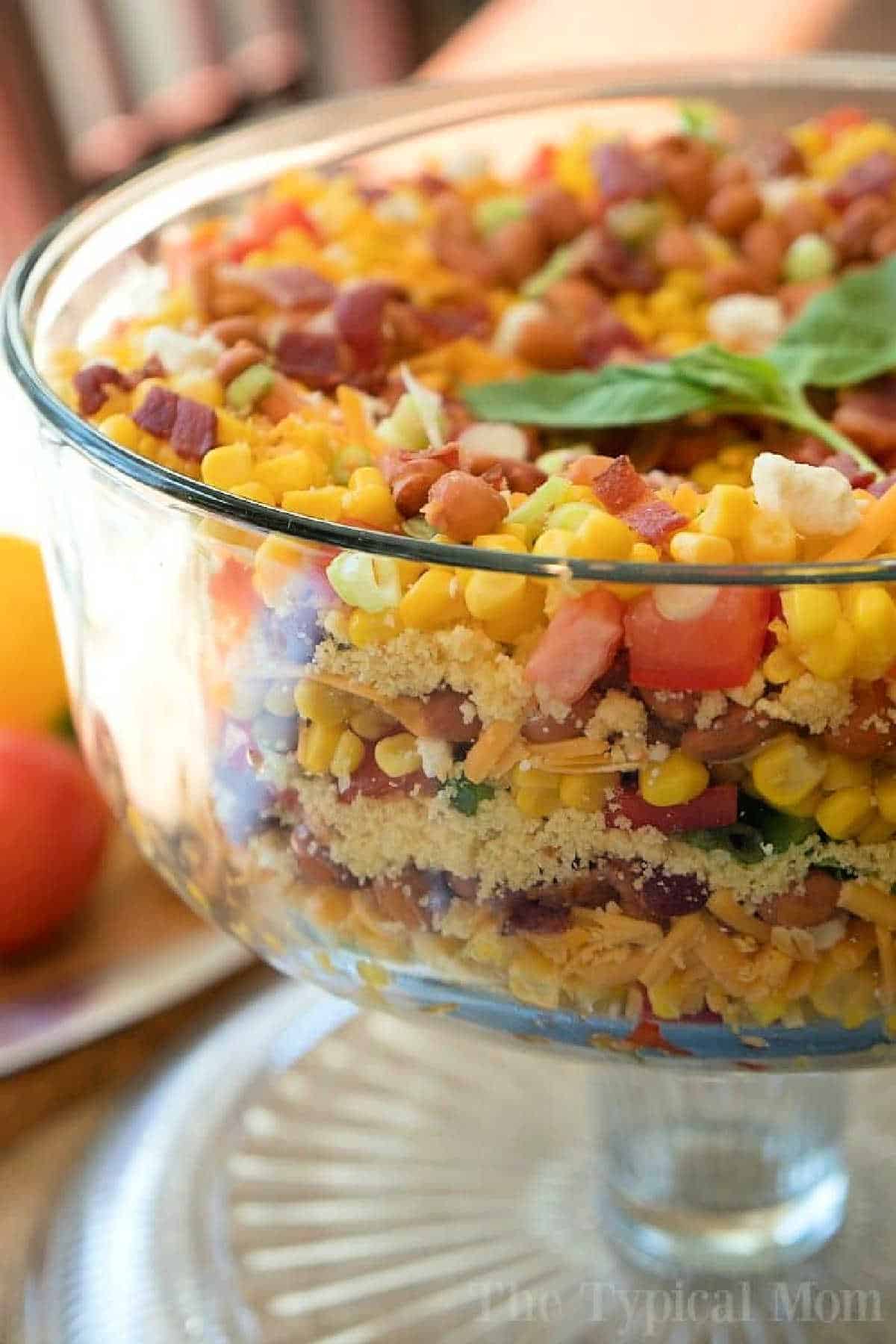 This easy layered cornbread salad recipe is a perfect side dish during the holidays or a barbecue! Layers of corn, cheese and more make it irresistible! #corn #salad #layered #cornbread #sidedish A glass bowl filled with layered Jiffy cornbread salad, including corn, cheese, bacon, beans, and chopped vegetables.