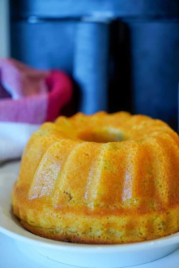 A golden brown air fryer cornbread bundt cake sits on a white plate, with a towel and dark background behind it.