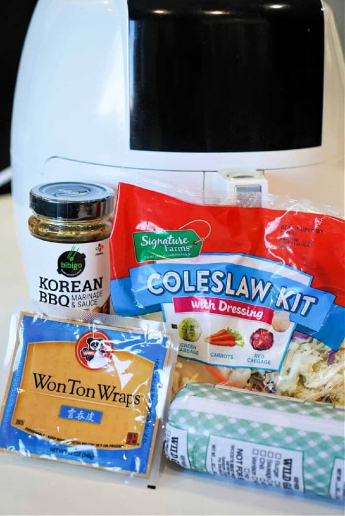 A jar of Korean BBQ sauce, coleslaw kit, wonton wraps, and ground pork in front of an air fryer&mdash;perfect for making flavorful air fryer won tons at home.
